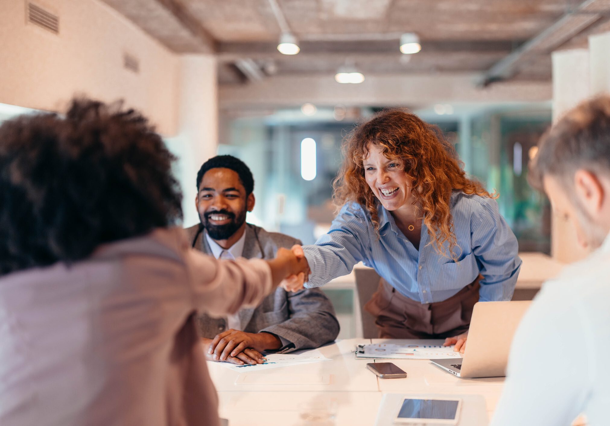 Two businesswomen shaking hands after successfully closing a deal during a productive meeting with colleagues in a modern office, celebrating teamwork and collaboration